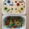 Insert sensoriel en bois avec fentes, trous picots et cercles de couleurs, avec pompons avec pompons, fleurs et picots dans un bac pour activité de tri et manipulation
