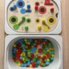 Insert sensoriel en bois avec fentes, trous picots et cercles de couleurs, avec pompons avec pompons, fleurs et picots dans un bac pour activité de tri et manipulation