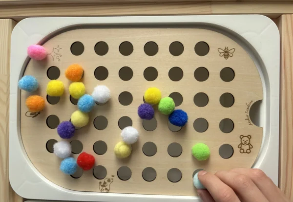 Insert sensoriel en bois avec trous pompons de 20mm avec pompons posés dessus avec main d'enfant qui joue