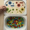 Insert sensoriel en bois avec fentes, trous picots et cercles de couleurs, avec pompons, fleurs et picots dans un bac pour activité de tri et manipulation avec la main d'un enfant