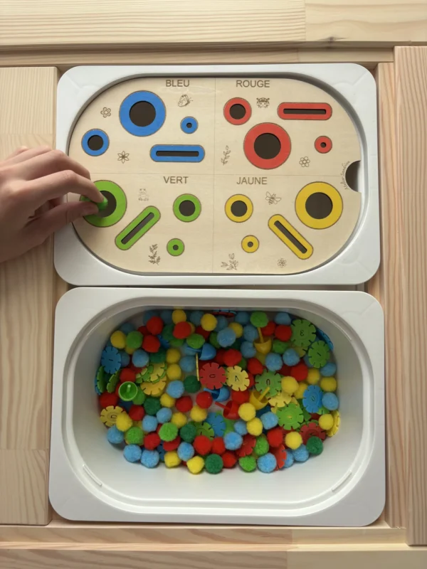 Insert sensoriel en bois avec fentes, trous picots et cercles de couleurs, avec pompons, fleurs et picots dans un bac pour activité de tri et manipulation avec la main d'un enfant