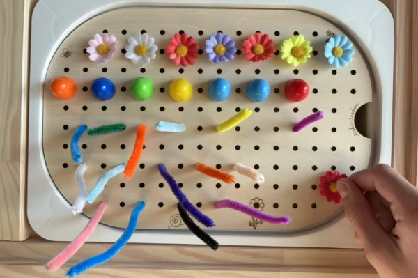 Insert sensoriel en bois avec trous picots plaque entière avec picots de couleurs disposés une couleur sur deux et fleurs et chenilles avec une main d'enfant qui joue