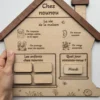 Main de femme tenant une maison des rituels en bois « Chez nounou » avec règles de vie, enfants présents et jour de la semaine.