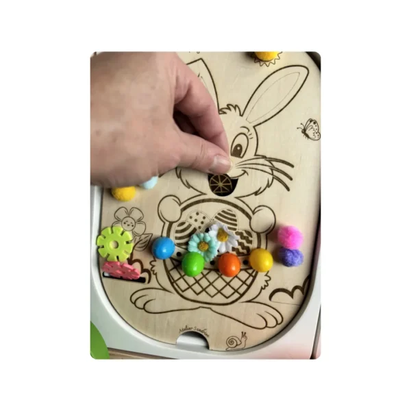 Main d’adulte manipulant un insert sensoriel en bois thème lapin de Pâques avec pompons colorés, favorisant la motricité fine et l’exploration sensorielle chez l’enfant.