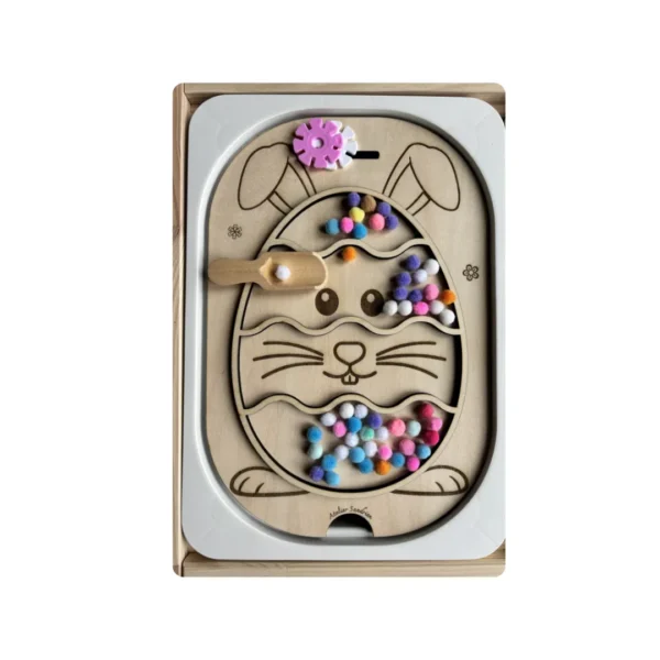 Insert sensoriel en bois thème Pâques en forme d’œuf avec lapin, utilisé pour remplir des pompons à l’aide d’une cuillère, activité favorisant la motricité fine, la coordination œil-main et l’autonomie des jeunes enfants.