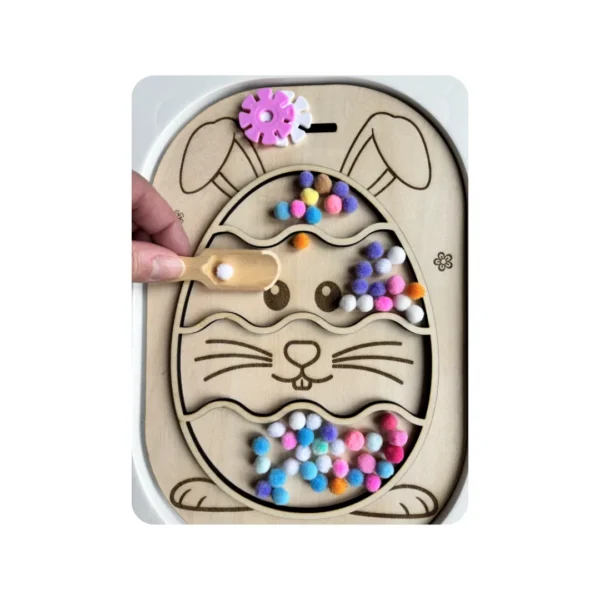 Insert sensoriel en bois thème Pâques en forme d’œuf avec lapin, utilisé pour remplir et manipuler des pompons colorés, activité favorisant la motricité fine, la coordination œil-main et l’autonomie des jeunes enfants.